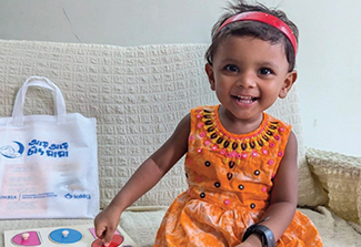 Photo of young Bangladeshi girl, smiling and wearing an orange dress and red headband, sitting on a couch with her hand on a wooden puzzle