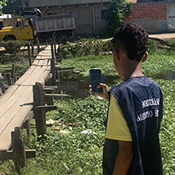 Teen wearing a yellow and blue shirt, holding a cellphone and taking a picture of a bridge