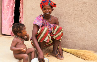 Woman in bright head wrap and clothing seated, smiling at camera on step next to naked toddler who is eating