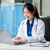 A young female doctor wearing a white lab coat and a stethescrope is working on a laptop in a medical office. She is smiling while interacting with a futuristic, holographic interface displaying medical and scientific icons. The background features blue hospital curtains and a clinical setting, with a tablet and coffee cup on the desk.