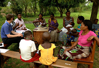 The photo shows a group of people, consisting of a couple of men and several women and children, sitting outside on wooden benches in a circle and engaging in discussion outside. Photo credit: Emily Mahoney/USAID