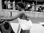 Young Ugandan woman, holding and looking at piece of fabric, with baby tied to her back, baby looks straight at camera 