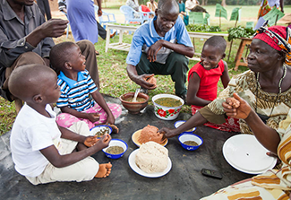 A group of people, including three adults and three children, sit together on the ground outdoors, sharing a meal.