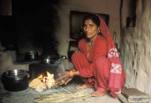 woman, barefoot, squats on the floor next to a cooking fire, dark smoke fills the air, no visible chimney or ventilation