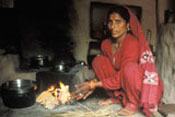 woman, barefoot, squats on the floor next to a cooking fire, dark smoke fills the air, no visible chimney or ventilation