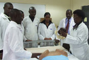 Group of medical students in white lab coats stands around hospital bed, woman leading discussion points to model she holds