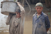 Two young men, mud on their shirts, stand in front of murky brown water, one holds large bucket on his shoulder 