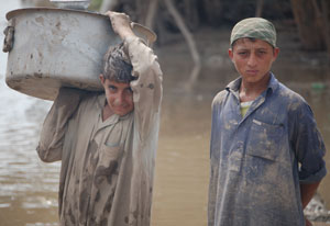 Two young men, mud on their shirts, stand in front of murky brown water, one holds large bucket on his shoulder 