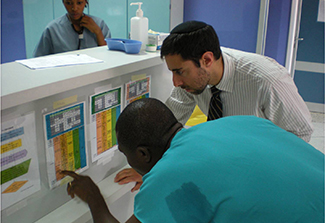 In this image, a MEPI student in Ghana, wearing blue scrubs, receives instructions from a teacher wearing a white patterned shirt and tie, on reading charts in the hospital. 