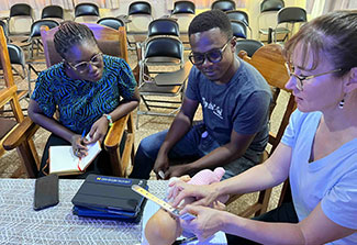 Cheryl Moyer sitting at a table with a woman and a man, demonstrating a clinical technique for trainees using a doll 