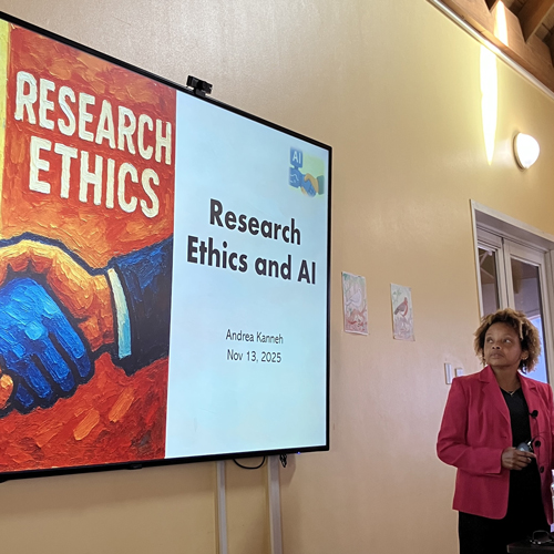 Woman wearing a pink blazer, looking at a larger screen that reads Research Ethics and AI