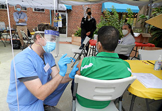 Dr. Michael Herce, wearing scrubs and protective mask, giving a shot in the arm to a man sitting in a chair 