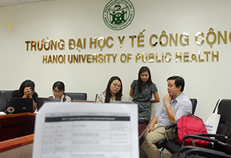 Group of five people seated in an office at Hanoi University of Public Health, with the university's name and logo displayed on the wall behind them; a paper document is visible in the foreground.