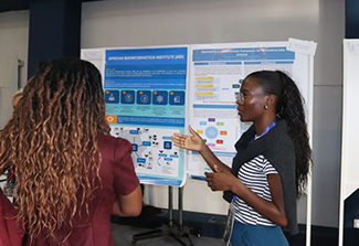 This photograph features an African woman, dressed in a striped top and blue pants, discussing her poster presentation to attendees of the DS-I African consortium meeting.