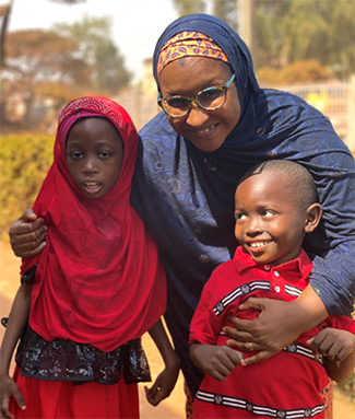 In this photo, Dr. Halima Bello-Manga, wearing a blue dress and headscarf, has her arms aroundtwo children, one boy wearing a red shirt with blue and white stripes and his sister, wearing a red dressand headscarf. The two children, both under age 10, are patients with sickle cell anemia.