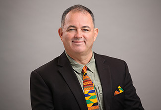 This page features a photo of Dr. Adel Driss wearing a black suit jacket, pale green shirt, and an  African-print tie with a matching pocket handkerchief.