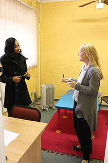 Two women stand talking in a room with bright yellow walls, one wearing a black coat and holding a small notebook, the other in a gray cardigan holding a pen and paper, with a red rug, blue table, and radiator visible in the background.