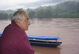 In the second photo on this page, Dr. Robert Gilman, wearing a maroon jacket and standing in profile, views the muddy waters of Puerto Ocopa in Peru as a blue boat passes.