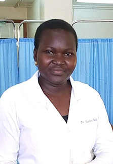 In this photo of Dr. Jackline Akello, she is seen at work in a clinic. She is wearing her white  doctor’s coat and stands before a blue curtained hospital divider.