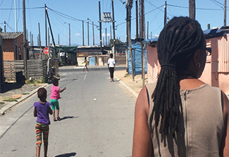 A person seen from behind, walks down a street in a township lined with small colorful houses and utility poles, while two children play nearby, one pointing toward a figure walking in the distance under a clear blue sky.