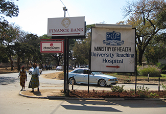 Street scene showing signs for Finance Bank, MoneyGram, and Ministry of Health University Teaching Hospital with a directional arrow, alongside a road where a car is driving and pedestrians are walking on the roadside.