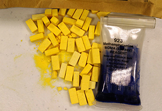 Photo of Methamphetamine scattered on a table next to a bag that reads 923 Methamphetamine MDMA Ecstasy REAGENT