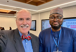 In this photo, Drs. Peter Kilmarx, wearing a navy suit, blue shirt and red bowtie, and Dr. Musa Kana, wearing an African fez and blue shirt, take a selfie together in a classroom at the 2024 UNGA Science Summit.