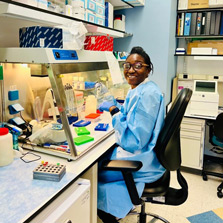 In this photo Dr. Vinie Kouamou wears blue protective gear as she sits at a lab desk and hoodworking with chemicals and other substances in various tubes