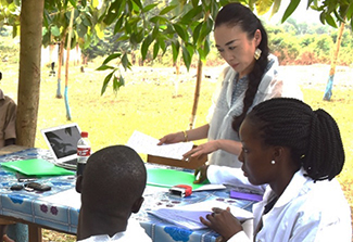 This photo shows Rie Yotsu (standing, right) reviewing paper work with mentees (seated). They are around a table set up outdoors and the all wear lab coats.