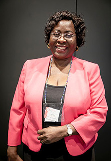 In this photo of Dr. Sarah Kiguli she is wearing a lanyard over her pink suit jacket with black  blouse and pants and she smiles broadly.