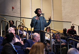 A female scientist, wearing a navy sweater, speaks into an audience microphone to ask Dr. Christian Happi a question.