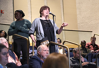 A female scientist, wearing a loose blue blazer over a black dress, speaks into an audience microphone to ask Dr. Christian Happi a question.