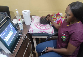 In this photo a healthcare worker, dressed in purple and blue scrubs, scans a child’s head with a probe as she conducts a Transcranial Doppler Ultrasound or TCD