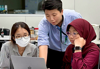 Three colleagues collaborate around a laptop in an office setting: a woman wearing a face mask sits on the left looking at the screen, a man in a blue shirt with a lanyard stands in the middle pointing toward the laptop, and a woman wearing a maroon hijab sits on the right resting her chin on her hand while looking at the screen.