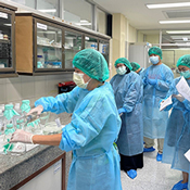 A group of healthcare workers wearing teal surgical gowns, caps, and masks gather in a hospital or lab room, with one person handling supplies at a counter while others hold clipboards and papers, some taking notes, in front of glass-fronted cabinets stocked with medical supplies.