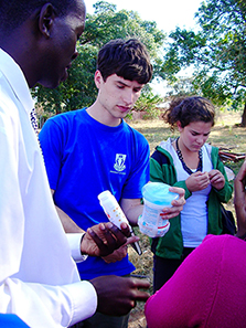 Under a Framework program, University of Virginia students present a water purification system in Limpopo, South Africa. The photo features a male student, wearing a bright blue t-shirt, and a female student, wearing a green jacket over a yellow t-shirt, talking with a local mentor wearing a white shirt and tie. 