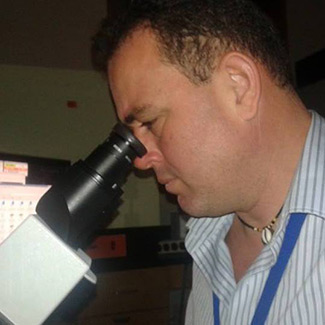 In the photo on this page, Dr. Adel Driss, who is wearing a white shirt with blue stripes, looks  into a microscope