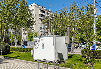 This photo shows a side view of air quality monitoring station in the Saburtalo district of Tbilisi, Georgia. The white rectangular structure is a few feet tall, wide and deep with an antenna-like metal pole on its roof