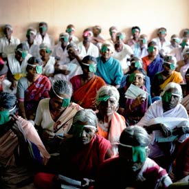 Many elderly Indian women in foreground and men in background seated on the floor, most wearing eye patches or bandages