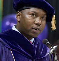 Dr Peter Cherutich speaks into a microphone wearing a purple doctoral tam and gown