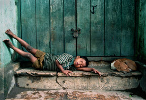 Young boy, barefoot and dirty, lies sleeping in doorway, appearing exhausted, dog curled up next to him on step