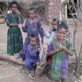 Five children look at and smile at the camera, playing in dirt courtyard, huts in background