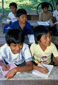 Two smiling boys write on paper pads on wooden desks in outdoor schoolhouse, more students in background do the same