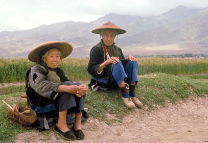 Two elderly women in china wearing Asian conical hats seated on the ground, farm fields and mountains in the background