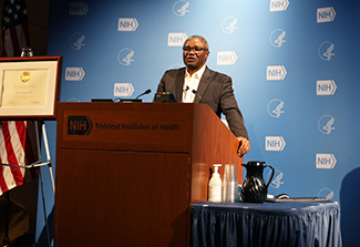 This photo shows Dr. Christian Happi, wearing a charcoal suit and a white, open collar shirt, as he stands at a podium on stage delivering the WALS lecture. 