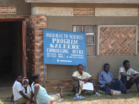 People seated on ground outside of building, sign on building reads Rakai health sciences program