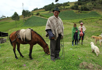 In lush green mountains of Colombia, a man looks at the camera holding the reigns of a horse, women and dogs in the background