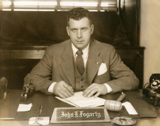 Black and white photograph of Congressman John E. Fogarty seated at a desk behind a nameplate with pen to paper