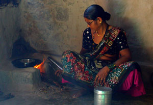 A woman seated on the floor next to a cookstove with a visible open flame adds wood to the fire