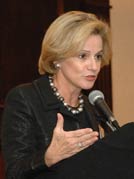 Woman speaks into a microphone at a podium, gestures with one hand